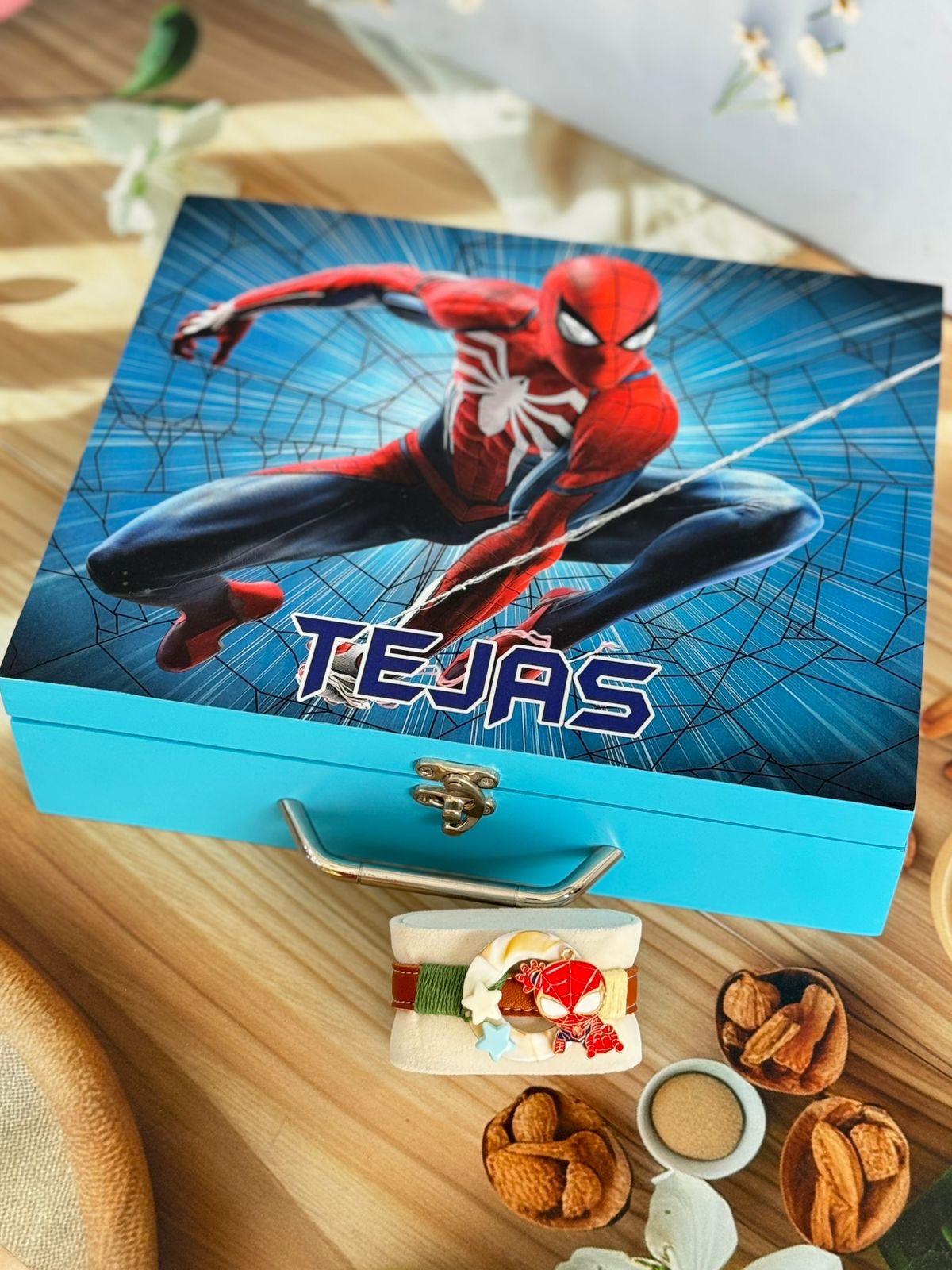 A personalized Spider-Man themed gift box with a bright turquoise base and a printed lid featuring Spider-Man on a blue webbed background, with the name 'TEJAS' printed on the lid. The box is styled on a wooden table with small food items in front.