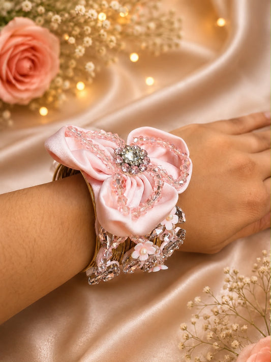 A close-up of a person's wrist wearing a wide pink satin bow and crystal-embellished bracelet, posed on draped champagne satin fabric with soft floral props and warm string lights.