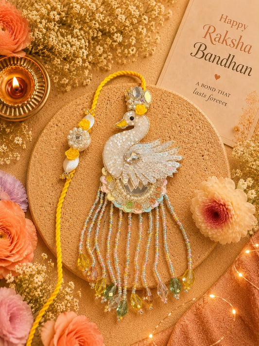 A decorative Rakhi with a white beaded peacock design and yellow tassel, displayed on a cork coaster with flowers, a lit diya, and a 'Happy Raksha Bandhan' card in a warm festive flat-lay arrangement.