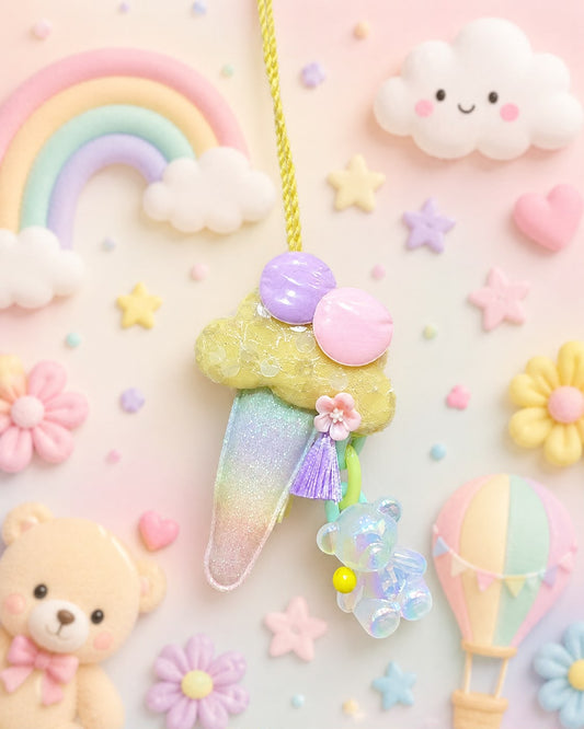 A pastel-themed hanging ornament featuring a glittery rainbow ice cream cone with a purple cherry on top, a small iridescent bear charm, and a purple tassel, displayed against a soft pink background decorated with a rainbow, clouds, stars, flowers, and a teddy bear.