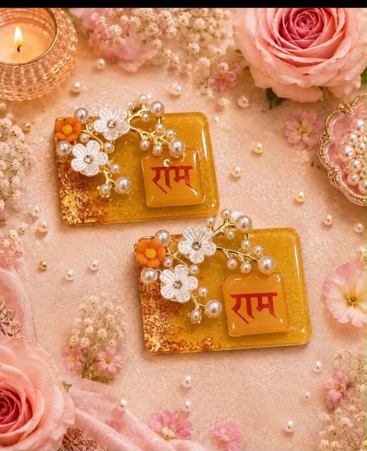Two rectangular gold-toned resin cards with the word 'Rām' in red Devanagari script, decorated with white and orange flowers and pearl-like beads, arranged on a pink floral surface with roses, baby's breath, and a lit candle.