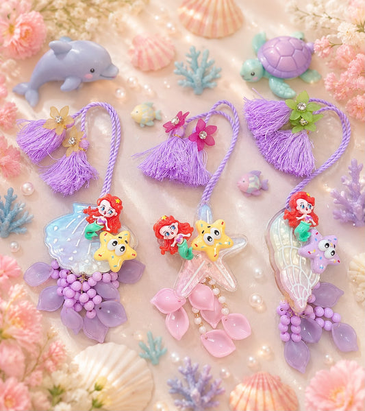 Three mermaid-themed resin keychains with purple tassels, arranged on a pastel pink background decorated with seashells, coral, pearls, and sea creatures.