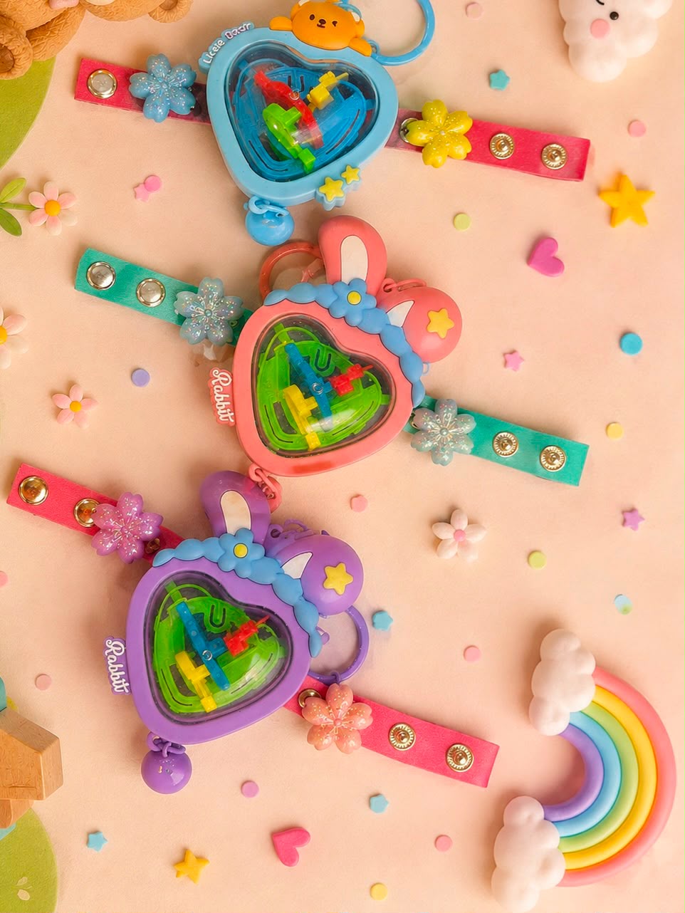 Three heart-shaped plastic toy keychains in pastel blue, pink, and purple, decorated with bunny ears, flowers, and stars, arranged on a pink background with small decorative flowers, stars, and a rainbow.
