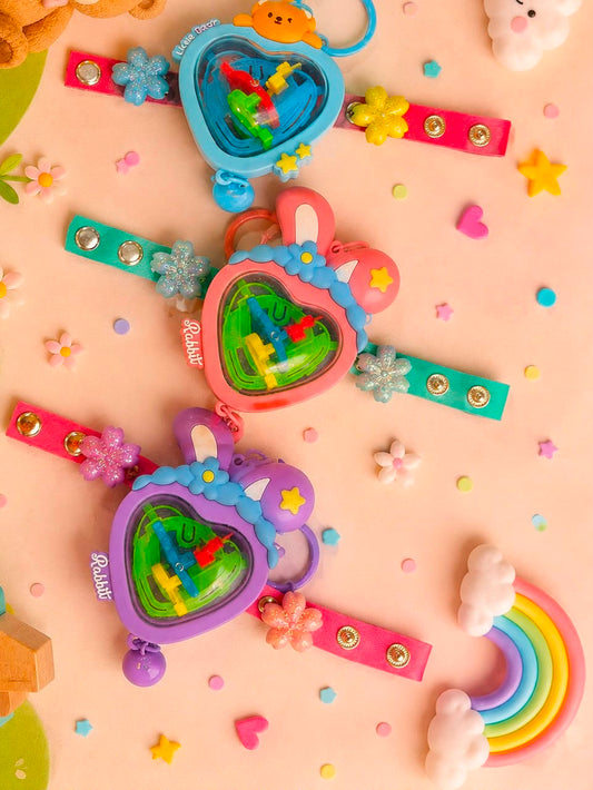 Three heart-shaped plastic toy keychains in pastel blue, pink, and purple, decorated with bunny ears, flowers, and stars, arranged on a pink background with small decorative flowers, stars, and a rainbow.