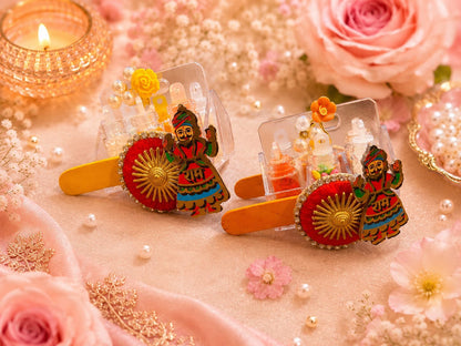 Two decorative hair clips styled with pink roses, pearls, and a lit diya. The clips feature miniature traditional Indian figures, red and gold circular motifs, and tiny doll figures inside clear compartments.