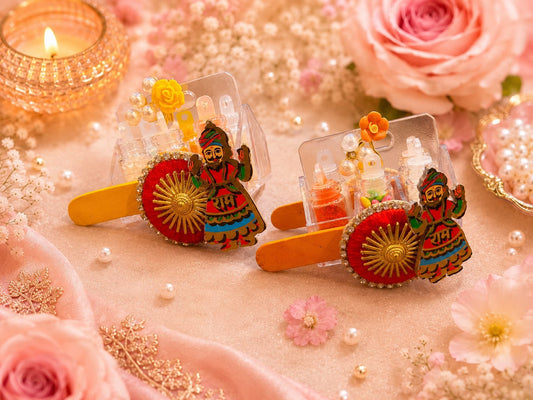 Two decorative hair clips styled with pink roses, pearls, and a lit diya. The clips feature miniature traditional Indian figures, red and gold circular motifs, and tiny doll figures inside clear compartments.