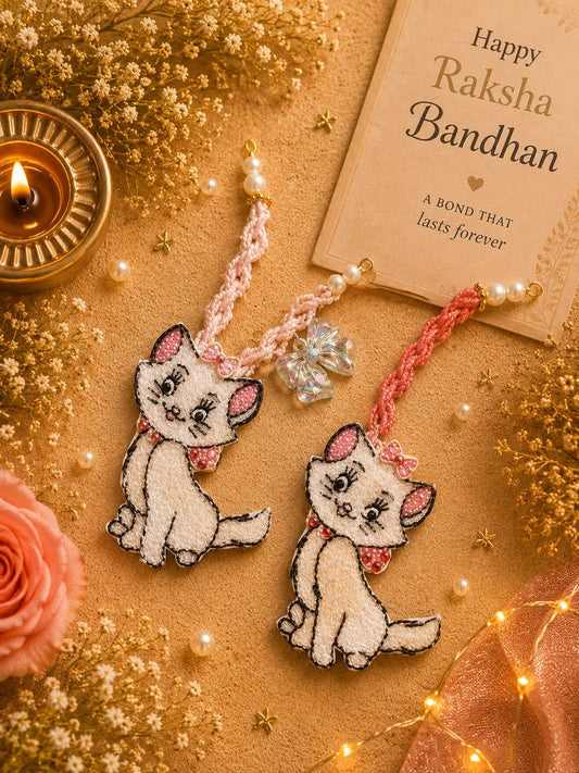 Two embroidered Rakhi-style cat ornaments with pink bows and pearl beads are displayed on a warm tan surface with dried flowers, a lit candle, string lights, and a card reading 'Happy Raksha Bandhan'.