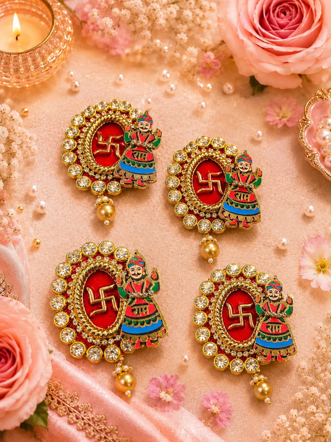 Four ornate gold-toned earrings arranged on a soft pink surface with decorative flowers, pearls, and a lit candle. Each earring has a red oval center with a gold swastika symbol and a colorful figure motif, surrounded by clear stones and dangling gold beads.