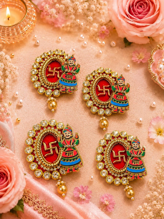 Four ornate gold-toned earrings arranged on a soft pink surface with decorative flowers, pearls, and a lit candle. Each earring has a red oval center with a gold swastika symbol and a colorful figure motif, surrounded by clear stones and dangling gold beads.