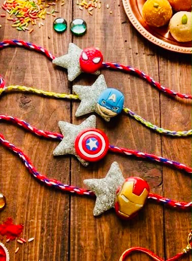Four colorful braided Rakhi strands laid out on a rustic wooden table, each decorated with a star-shaped bead and a round character bead featuring Marvel superheroes: Spider-Man, Captain America, Captain America, and Iron Man. A plate of sweets is partially visible in the top right corner.