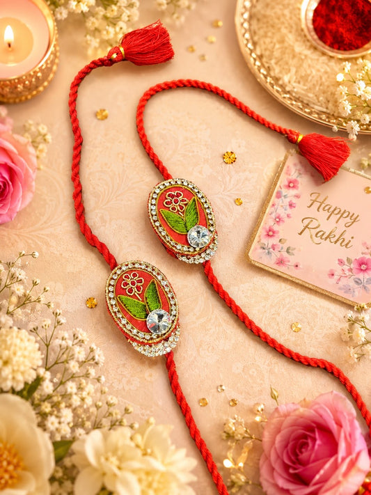 Two red Rakhi cords with oval decorated medallions, displayed on a beige surface with flowers, a lit candle, gold decorative elements, and a small 'Happy Rakhi' card.