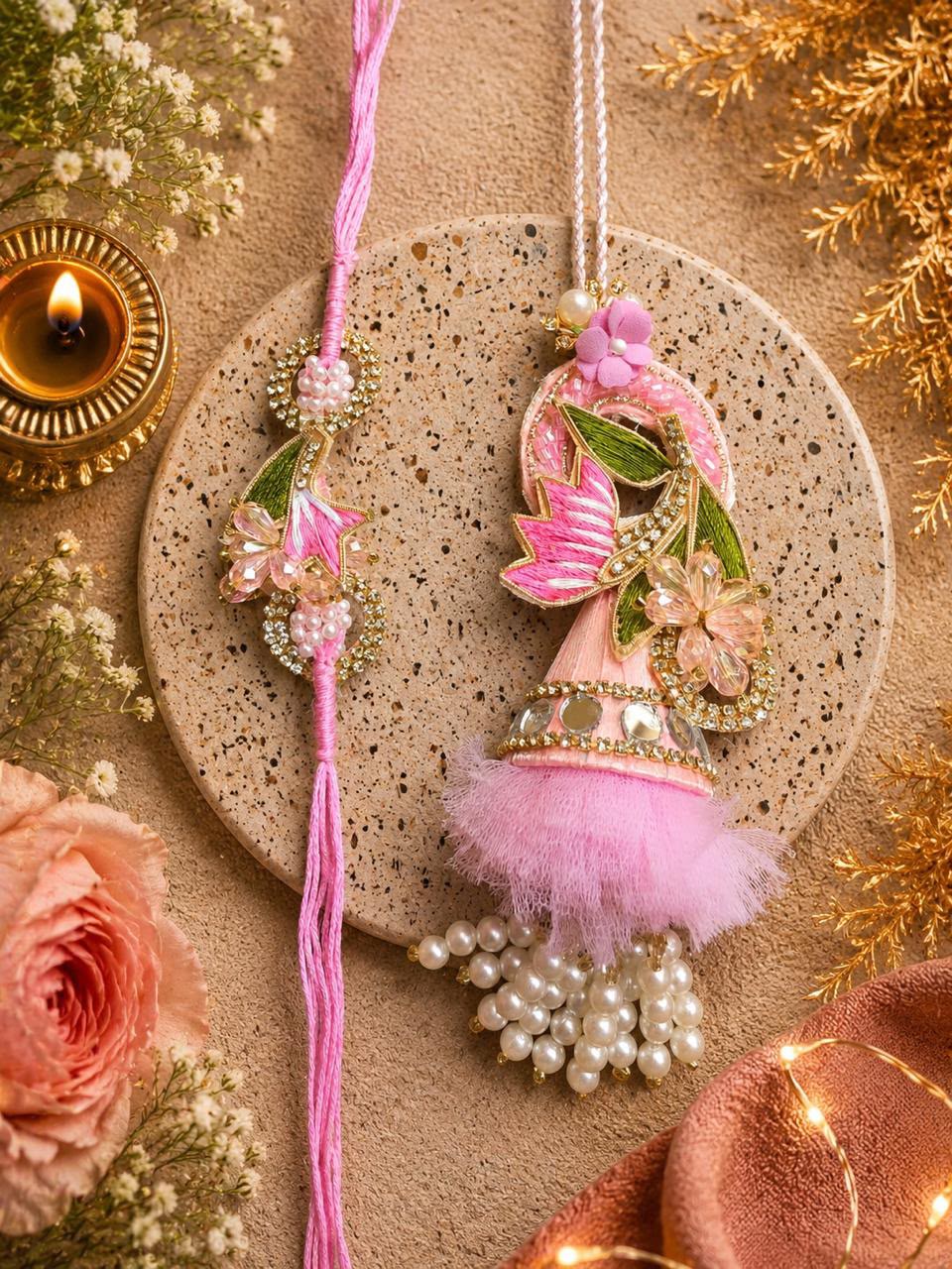A decorative pink Rakhi with a tassel and pearl bead garland is displayed alongside a matching pink thread Rakhi on a speckled stone plate, surrounded by floral props, a lit diya, and warm holiday decorations.