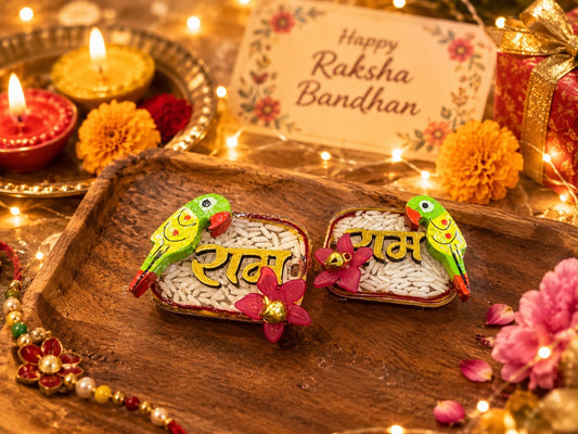 Two decorated rectangular plates filled with white rice and topped with colorful parrot figurines and pink flowers, placed on a wooden tray in a festive Diwali setting with candles, marigold flowers, and a card reading 'Happy Raksha Bandhan'.