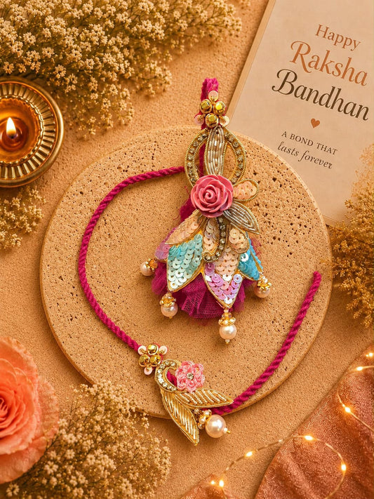 A decorative Rakhi with a pink cord and ornate gold-toned pendant featuring a pink rose, sequined leaf, and pearl beads, displayed on a round cork coaster with a lit diya, dried flowers, and a Rakhi card in the background.