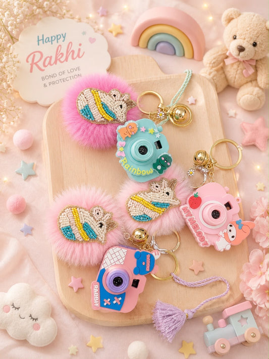 A styled flat-lay of four cute keychains and hair clips arranged on a wooden tray, surrounded by pastel props including a teddy bear, rainbow, clouds, stars, and a 'Happy Rakhi' card. The products feature pink fluffy pom-poms, embroidered hedgehogs, and camera charms in pastel rainbow colors with gold-tone hardware.