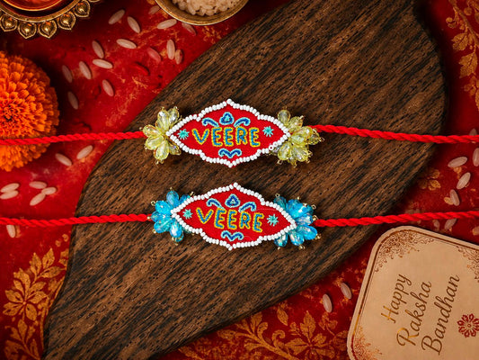 Two handmade Rakhi bracelets with 'VEERE' embroidered in the center, displayed on a dark wooden spoon over a red patterned fabric background with festive decorative elements.