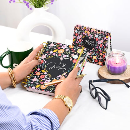 Person holding a floral planner with 'my journey' text, surrounded by a mug, glasses, and a candle on a table.