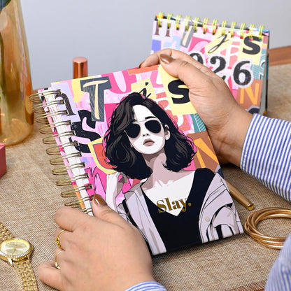 Person holding a colorful notebook with an illustration of a person wearing sunglasses and the word 'Slay'.